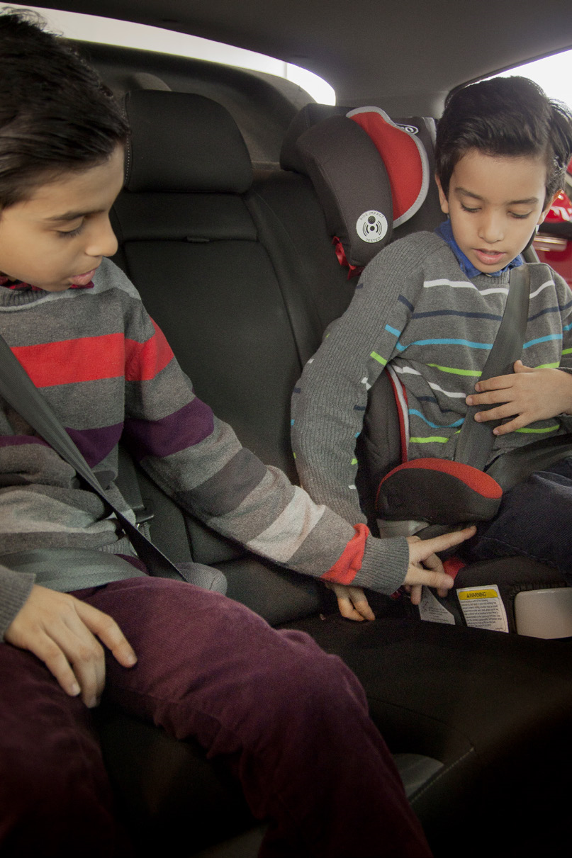 Two children sitting next to each other on booster seats, buckled into the seat with the seat belt. One booster seat is a high-back booster seat with a head rest.