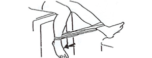 Woman sitting on a chair, with a rubber band stretched between both feet. She has one leg pointed straight outward, and is pulling the other foot in towards herself.
